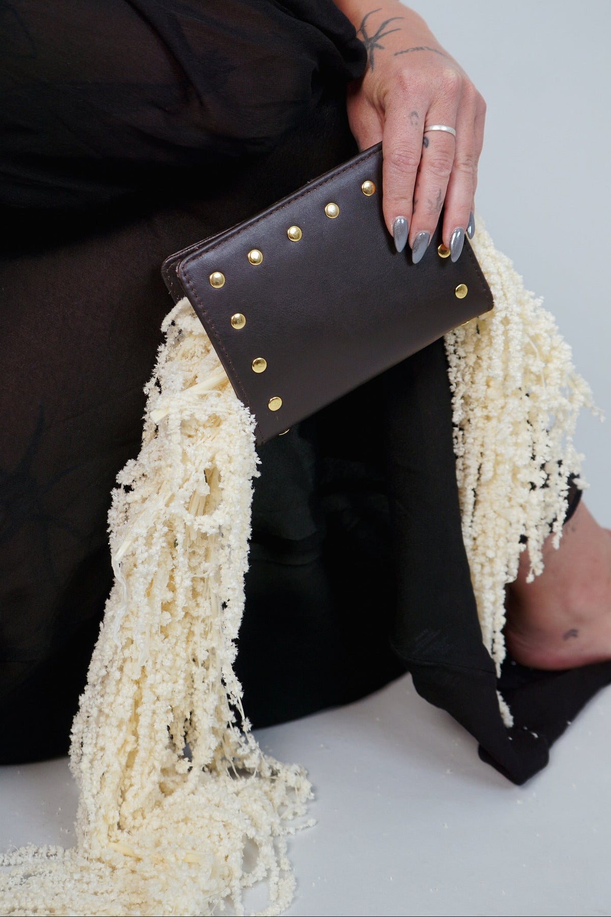 Hand holding the dark brown Amaretto passport case with gold studs against a cream fringed shawl