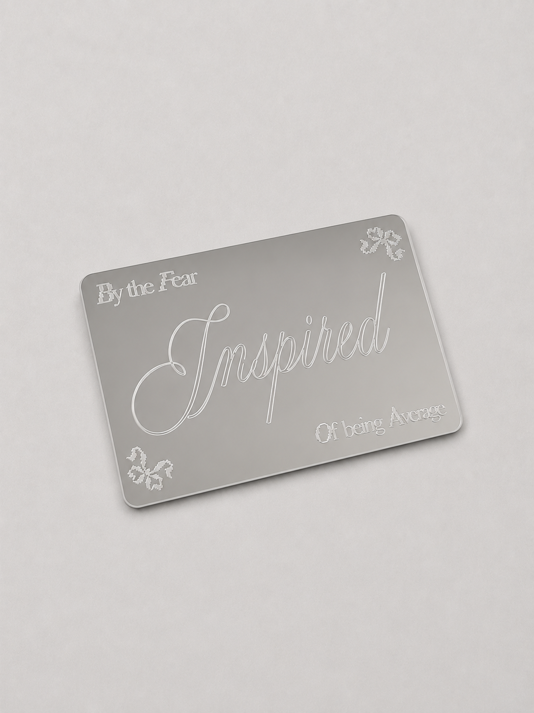 A silver rectangular mirror card with frosted white text reading 'By the Fear', 'Inspired', and 'Of being Average', plus small decorative floral motifs, shown on a light gray background.