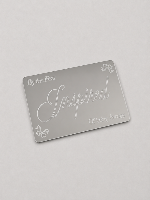 A silver rectangular mirror card with frosted white text reading 'By the Fear', 'Inspired', and 'Of being Average', plus small decorative floral motifs, shown on a light gray background.
