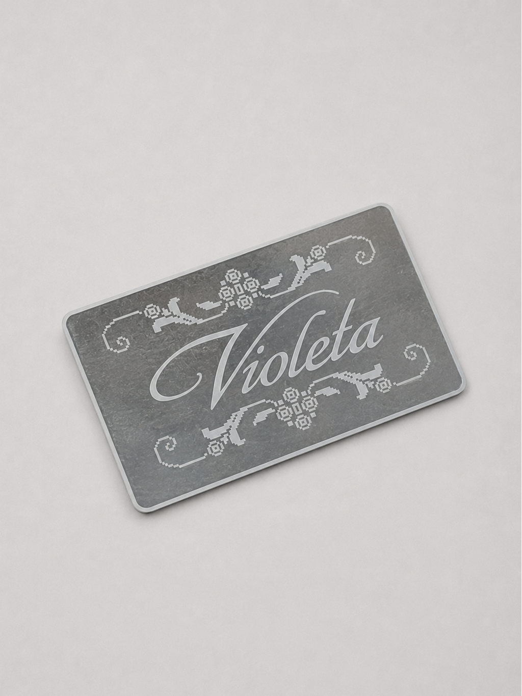 A rectangular mirrom card with rounded corners, featuring the word 'Violeta' in white cursive script and white decorative flourishes, photographed on a light gray background.