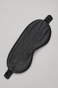 A black satin sleep mask with an embossed script logo on the front, displayed on a neutral light gray background.