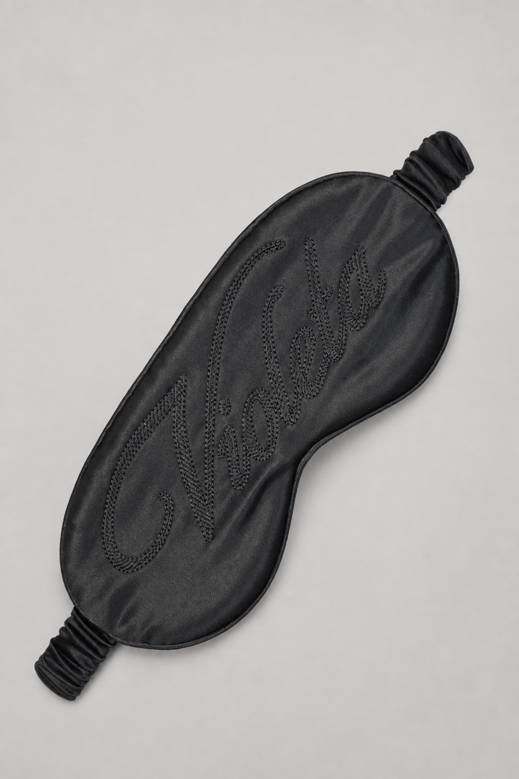A black satin sleep mask with an embossed script logo on the front, displayed on a neutral light gray background.
