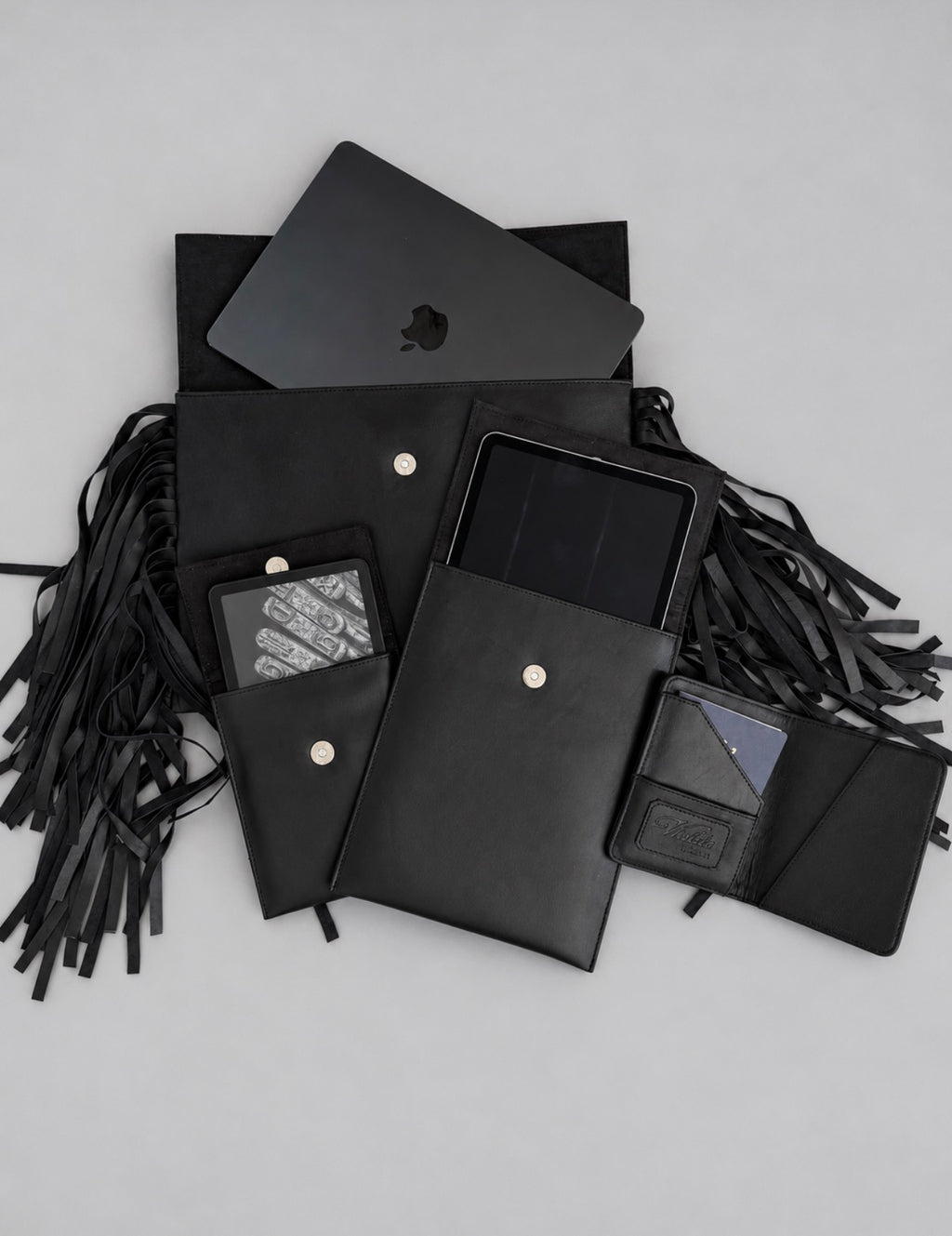 Violeta Shalom collection flat lay: fringe laptop case with a MacBook, iPad case, Kindle case and open passport case in black leather
