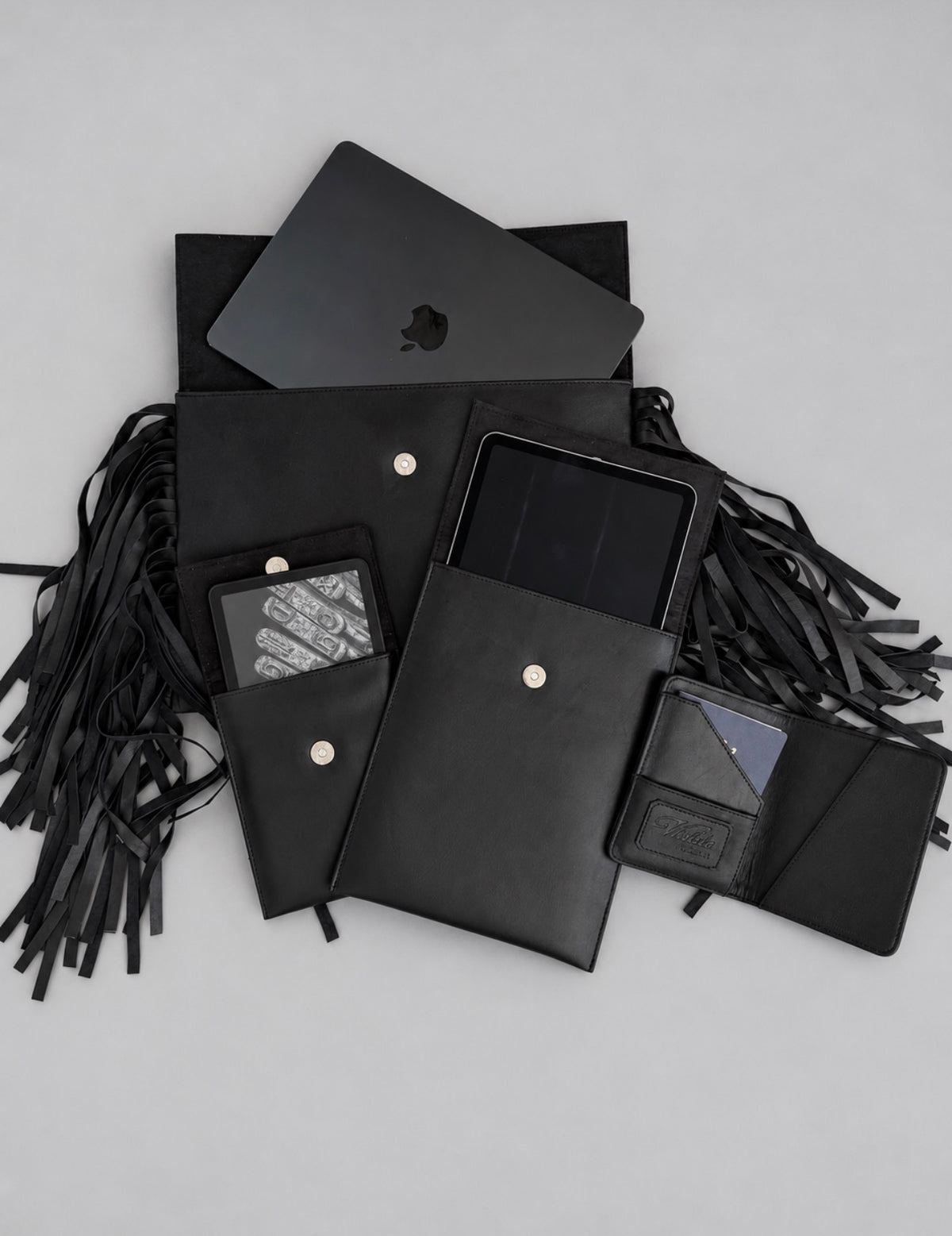 Violeta Shalom collection flat lay: fringe laptop case with a MacBook, iPad case, Kindle case and open passport case in black leather