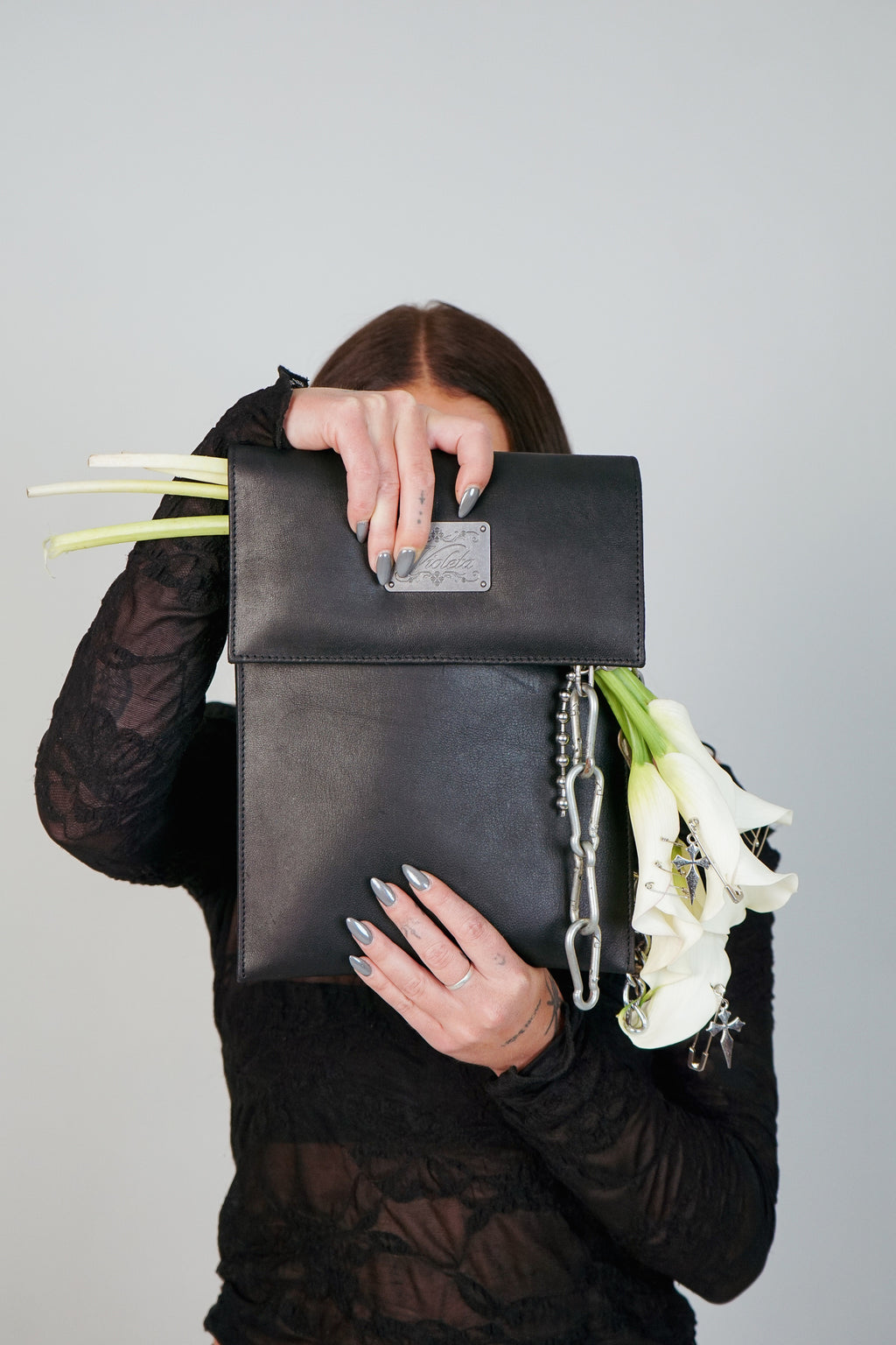 Woman in a black lace top holding the black leather Shalom iPad case up to her face, with white calla lilies and a silver chain